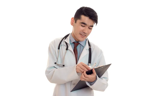 性格开朗的年轻黑发人医生穿白色的制服与听诊器在使被隔绝在工作室的笔记 — 图库照片