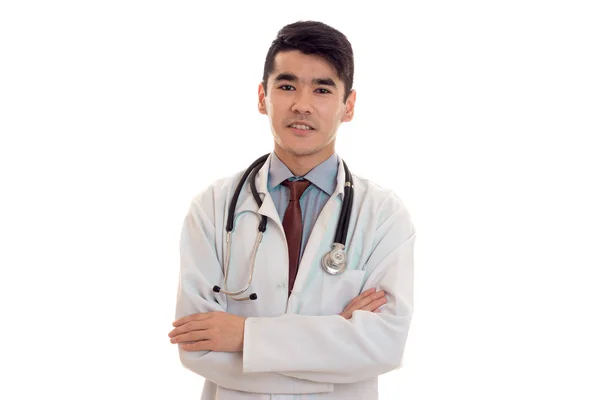 Beau jeune homme brunette médecin en uniforme blanc avec stéthoscope et mains croisées isolé en studio — Photo