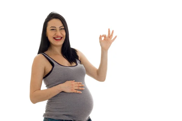 Happy young brunette pregnant girl with hand on her belly showing OK isolated on white background — Stock Photo, Image