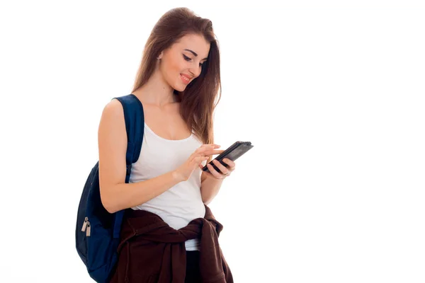 Fröhliches intelligentes Studentenmädchen in brauner Sportkleidung mit Rucksack auf den Schultern, das auf sein Handy blickt und isoliert auf weißem Hintergrund lächelt — Stockfoto