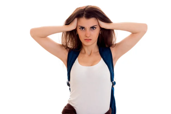 Anfrar menina estudante inteligente em roupas esportivas marrom com mochila em seus ombros olhando para a câmera com as mãos na cabeça isolada no fundo branco — Fotografia de Stock
