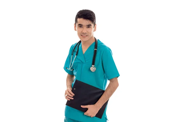 Primer plano retrato de joven guapo morena hombre médico en uniforme azul con estetoscopio y tabla en las manos mirando a la cámara y sonriendo aislado sobre fondo blanco —  Fotos de Stock