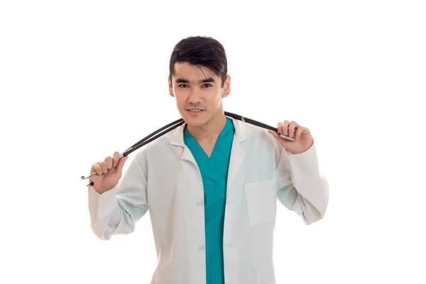 Retrato de jovem bonita morena homem médico em uniforme branco com estetoscópio olhando para a câmera isolada no fundo vazio — Fotografia de Stock