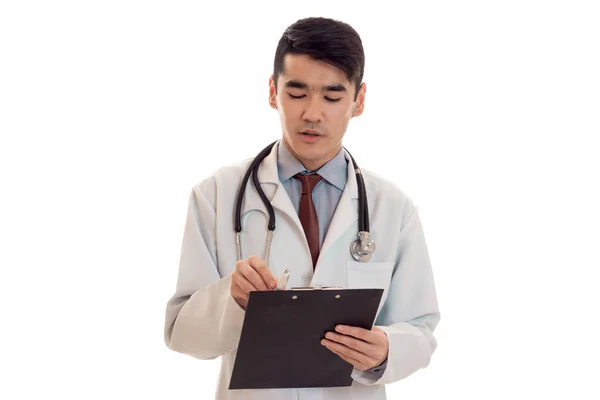 Retrato de joven guapo morena hombre médico en uniforme blanco posando en cámara con estetoscopio en su cuello hacer notas aisladas sobre fondo vacío —  Fotos de Stock