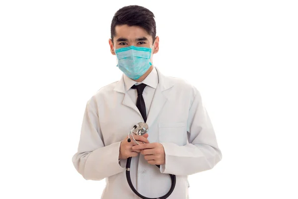 Portrait de jeune homme brune gai médecin en uniforme blanc et masque bleu avec stéthoscope sur son cou regarde la caméra isolée sur fond vide — Photo