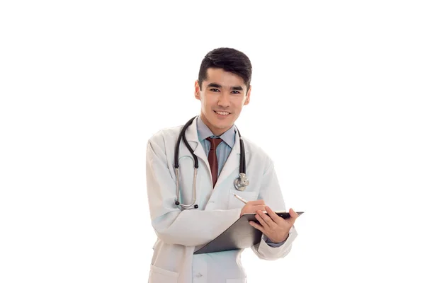 Joven médico alegre mantiene un estetoscopio en los hombros y el tablero en la mano aislado sobre fondo blanco — Foto de Stock
