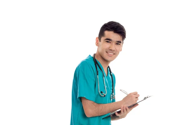 Neşeli esmer delikanlı doktor mavi üniforma onun omuzlarında stetoskop ile yapmak notlar ve beyaz arka plan üzerinde izole kamera üzerinde gülümseyen — Stok fotoğraf