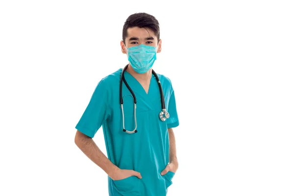 Joven guapo morena hombre médico en uniforme y máscara con estetoscopio en sus hombros mirando a la cámara aislada sobre fondo blanco —  Fotos de Stock