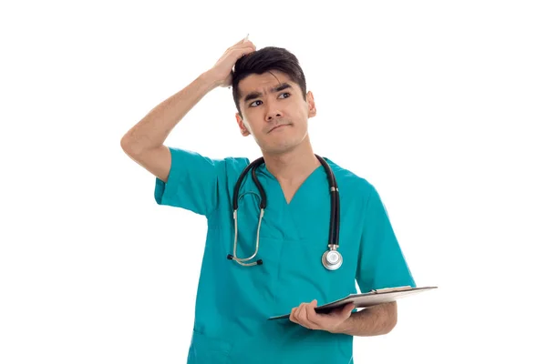 Young funny brunette man doctor in blue uniform with stethoscope on his shoulders make notes and thinking isolated on white background — Stock Photo, Image