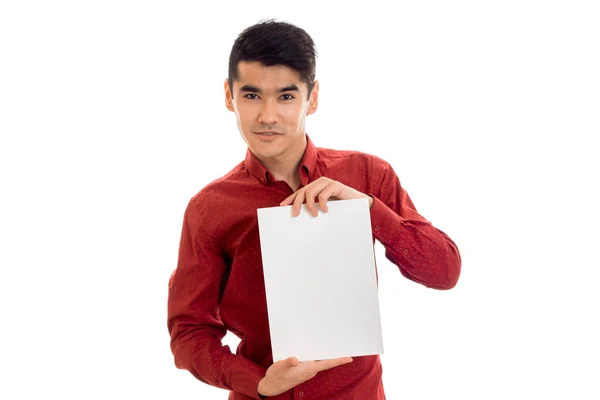 Youn elegante morena modelo masculino em camisa vermelha posando com cartaz vazio em suas mãos e olhando para a câmera isolada no fundo branco — Fotografia de Stock