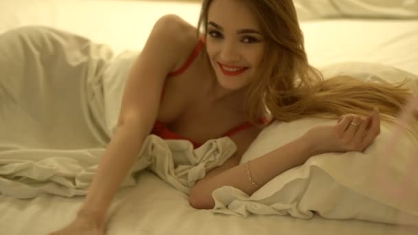 自宅の白いベッドでポーズをとって赤いランジェリーの陽気な若いブロンドの女性 — ストック動画