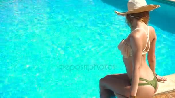 Jovencita sexual con sombrero de paja y traje de baño seductor se sienta junto a la piscina azul — Vídeo de stock