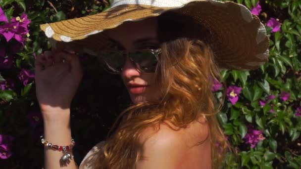 Giovane ragazza bionda in occhiali da sole e cappello di paglia in posa nel giardino fiorito — Video Stock