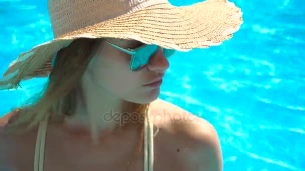 A young girl wearing a hat and glasses stands near the pool and looks towards — стоковое видео