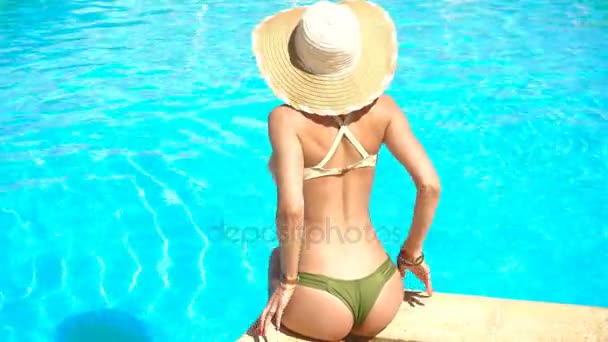Jovem menina em terno de banho sexy e chapéu sentado à beira da piscina e corrige calcinha — Vídeo de Stock
