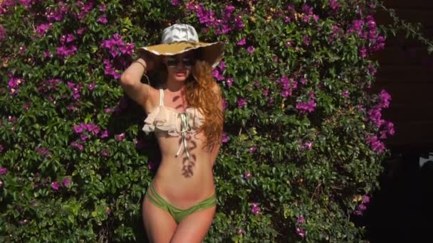 Красивая молодая блондинка в соломенной шляпе и сексуальное бикини позирует в цветущем саду с цветами — стоковое видео