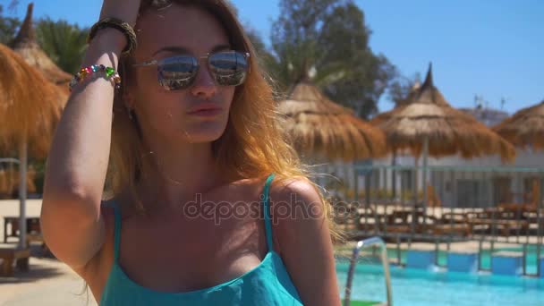 Joven hermosa chica en gafas vale la pena junto a la piscina en el verano y mantiene una mano cerca del cabello en cámara lenta de cerca — Vídeos de Stock