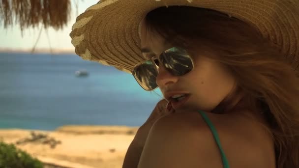 Молода сексуальна дівчина в окулярах і капелюсі позує на камеру біля моря, а вітер дме у повільному русі крупним планом — стокове відео