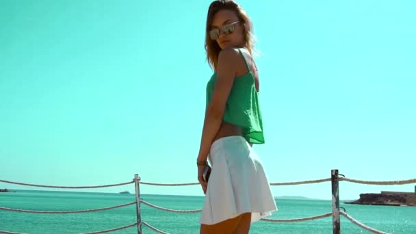 Mooie meisje in glazen en witte rok staat op een pier in de buurt van de zee en kijkt naar de camera in slow motion — Stockvideo