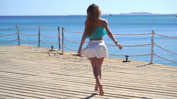 Молодая девушка в белой юбке и с длинными волосами бежит по пирсу в море в замедленной съемке — стоковое видео