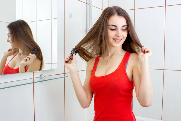 Vacker ung flicka i en röd väst med långt hår står mittemot spegeln — Stockfoto