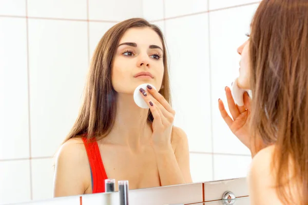 Jovem menina bonita de pé ao lado de um espelho e limpa o rosto com um disco de algodão — Fotografia de Stock