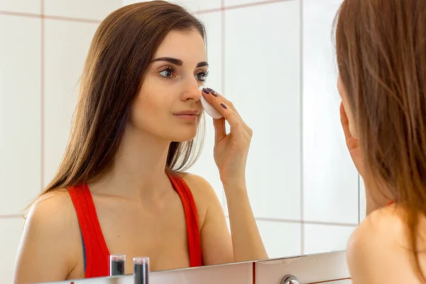 Красива молода дівчина в червоній сорочці стоїть перед дзеркалом і протирає обличчя ватним диском — стокове фото