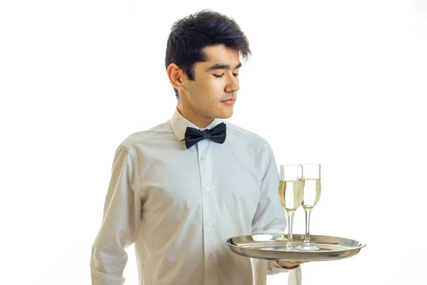 Encantador jovem garçom em uma camisa branca e com uma borboleta segurando uma bandeja com dois copos de champanhe — Fotografia de Stock