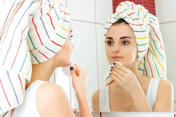 Милая молодая девушка с полотенцем на волосах смотрит в зеркало и массирует лицо с хлопковым диском — стоковое фото