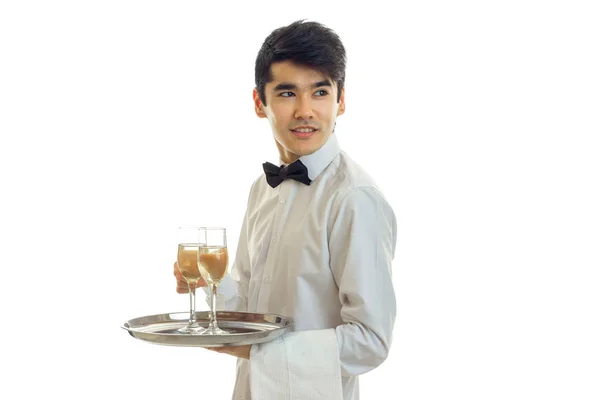 Przystojny, młody kelner kokarda krawat i koszula wygląda odległości i utrzymuje kieliszki do szampana na tacy, który — Zdjęcie stockowe