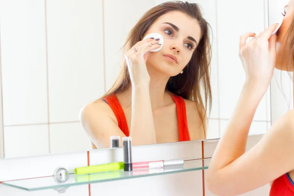 Mooie jonge meisje staat voor een spiegel en wrijft uw gezicht lotion met een katoen schijf — Stockfoto