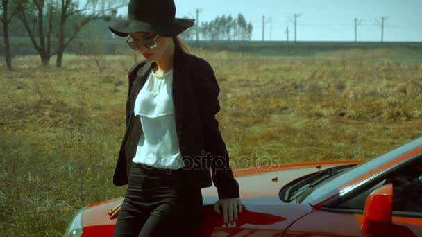 Dame in zonnebril en hoed met brede rand staat met een rode auto — Stockvideo