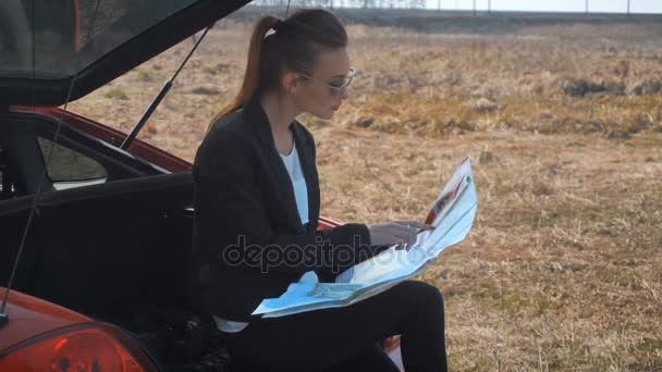 Девушка сидит в багажнике машины и исследует карту — стоковое видео