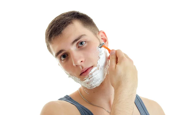 Joven con espuma en la cara mira a la cámara y se afeita la máquina de barba — Foto de Stock