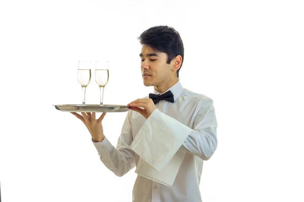 Przystojny, młody kelner z czarnymi włosami stoi prosto odwraca wzrok i posiada tackę z dwie szklanki — Zdjęcie stockowe