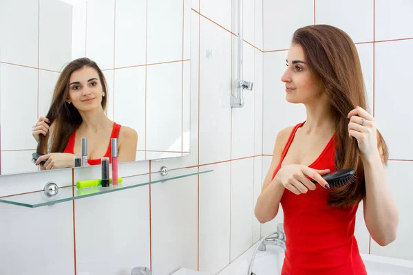 Ung vacker flicka i en röd väst kammar sitt hår framför en spegel — Stockfoto
