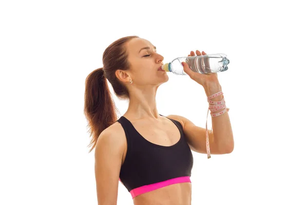Unga fitness girl står i sidled i sport topp och dricksvatten från en flaska — Stockfoto