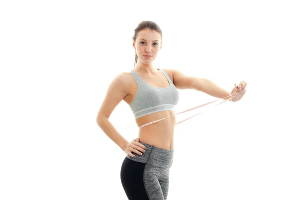 Attractive young girl in grey sports top looks straight and pulls at the waist measuring tape — Stock Photo, Image
