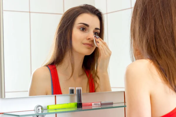 Нежная молодая девушка, стоящая в ванной перед зеркалом и массирует лицо лосьоном — стоковое фото