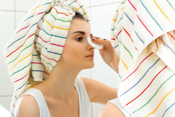 Jong meisje met handdoek op het haar in de spiegel kijkt en zachtjes veegt haar gezicht met een katoen schijf — Stockfoto