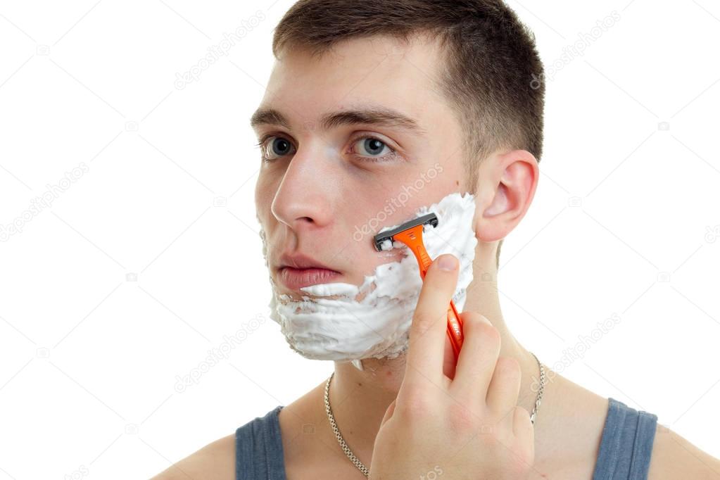 Portrait of a handsome young guy with foam on his face who carefully shaves his beard machine close-up