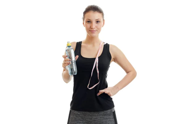 Cute young fitness girl in black shirt looks into the camera and keep the measuring tape and water at hand — Stock Photo, Image