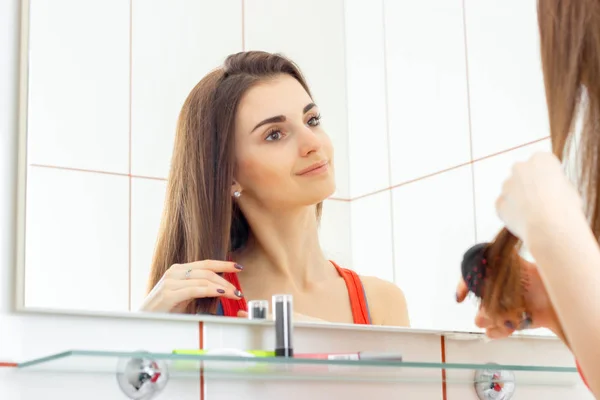 Mignonne jeune fille souriante se tient devant un miroir et peigner ses cheveux longs — Photo