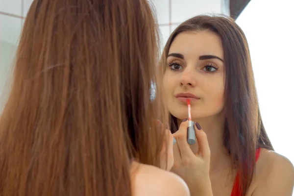 Привлекательная молодая девушка с длинными волосами стоит перед зеркалом и красит губы — стоковое фото