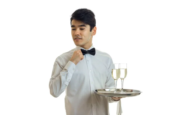 Czarujący przystojny młody kelner z czarnymi włosami dostosowuje jego muszką i ręka trzyma tacę z dwoma okularami — Zdjęcie stockowe