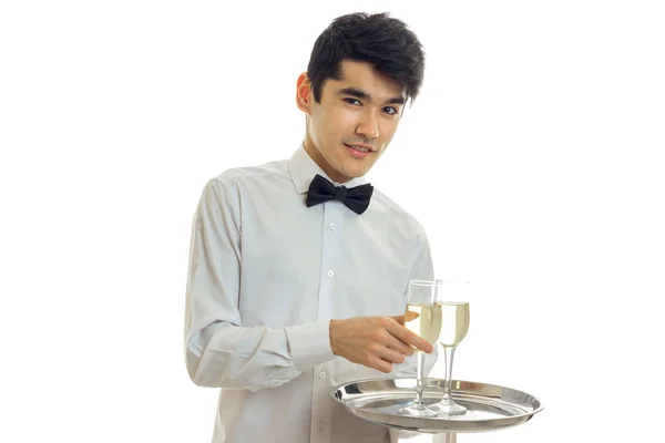 Очаровательный молодой официант улыбается, смотрит в камеру и держит поднос с бокалами шампанского — стоковое фото