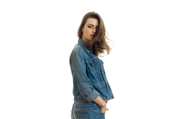 Giovane bruna carina con bei capelli si erge lateralmente in un abito jeans e guardando la fotocamera — Foto Stock