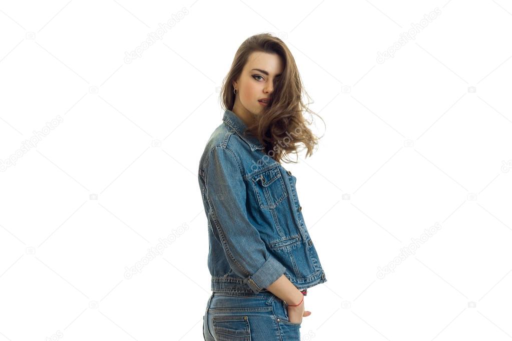 young cute brunette with beautiful hair stands sideways in a jeans suit and looking at camera