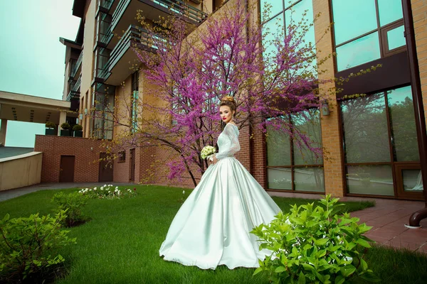 Süße junge blonde Braut im Hochzeitskleid schaut draußen vor dem Sakura-Baum in die Kamera — Stockfoto