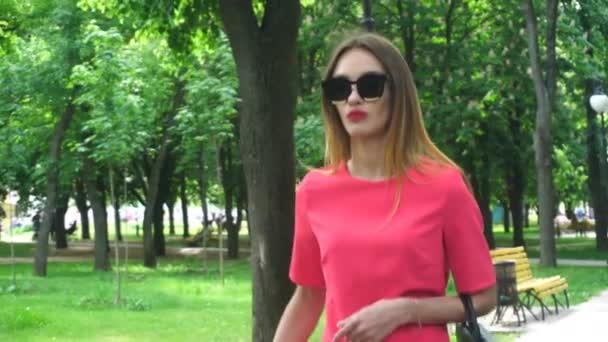 Mujer joven en gafas de sol y un traje rojo camina en el parque — Vídeos de Stock